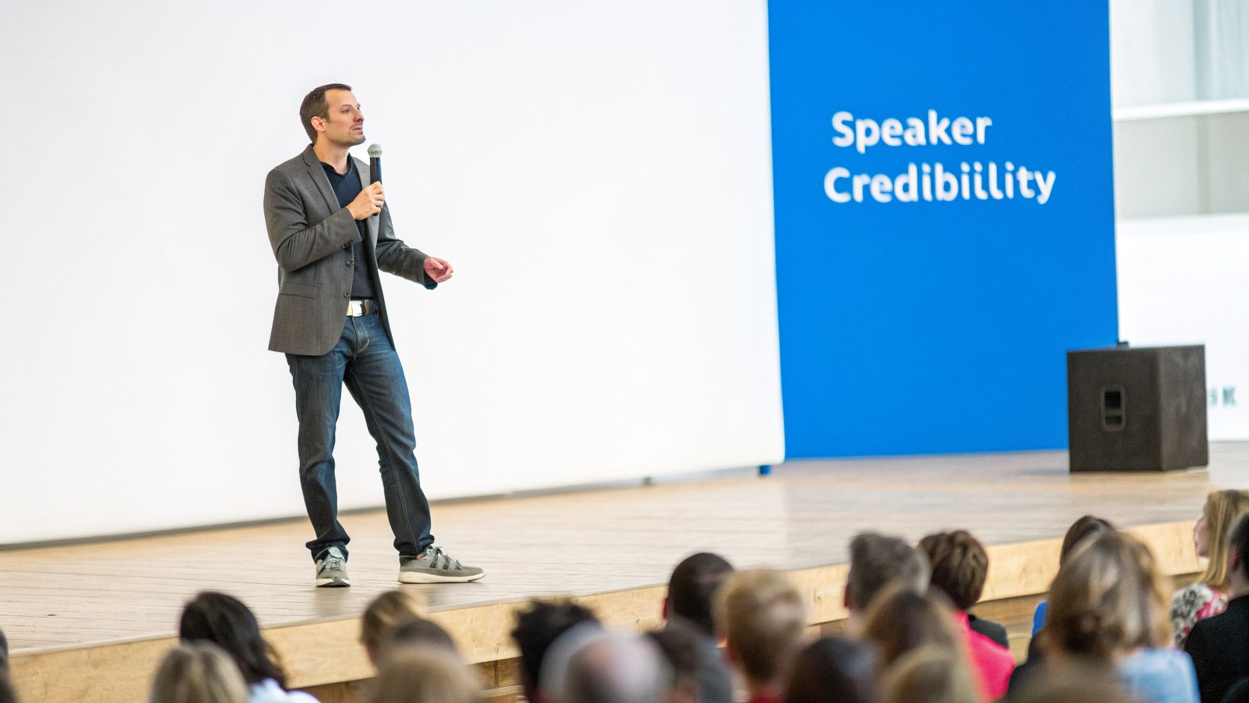 A man in a grey suit jacket and jeans speaks on stage, holding a microphone, with 'Speaker Credibility' on a blue backdrop.