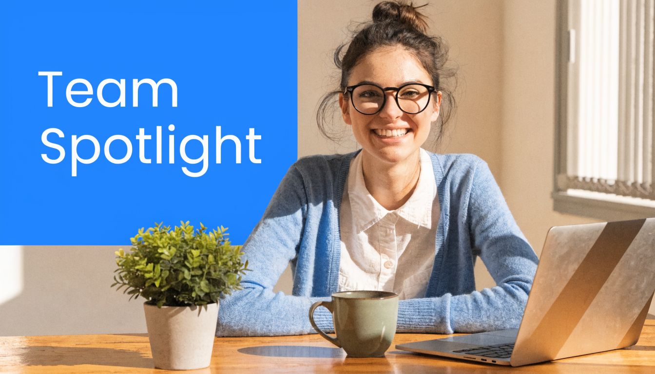 A smiling woman with glasses working on her laptop at a desk with a plant and coffee.