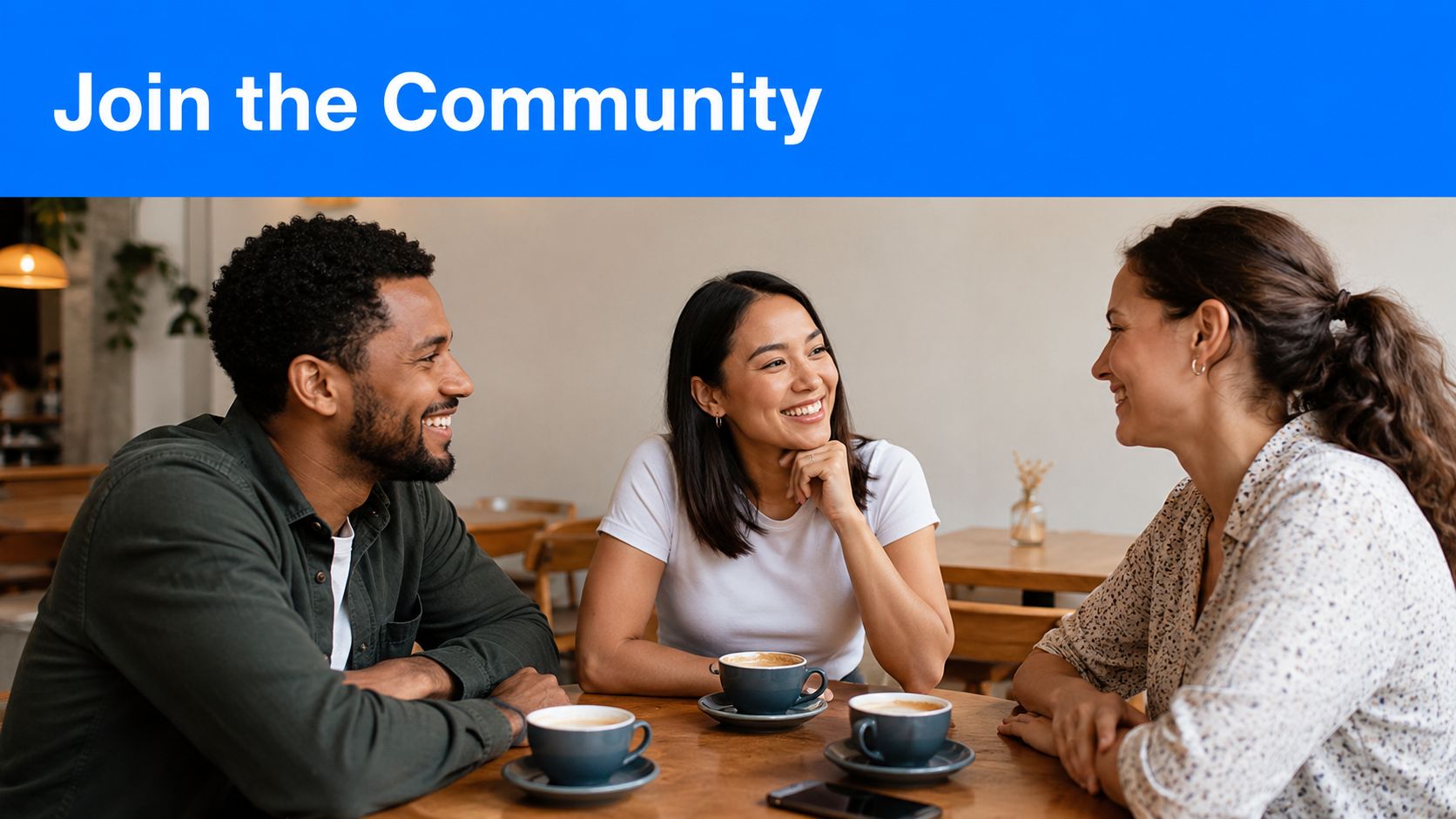 Three diverse friends sitting at a cafe table smiling, talking, and drinking coffee together.